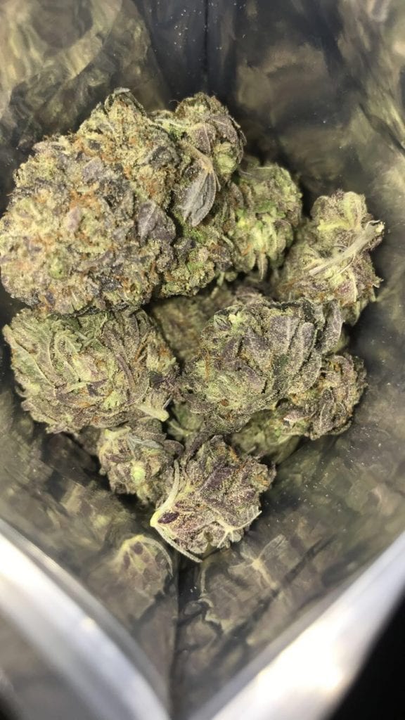 On the inside of a silver aluminum bag, there are a variety of different size purple nuggets of cannabis. They have dark purple coating them with a hint of green in the background.