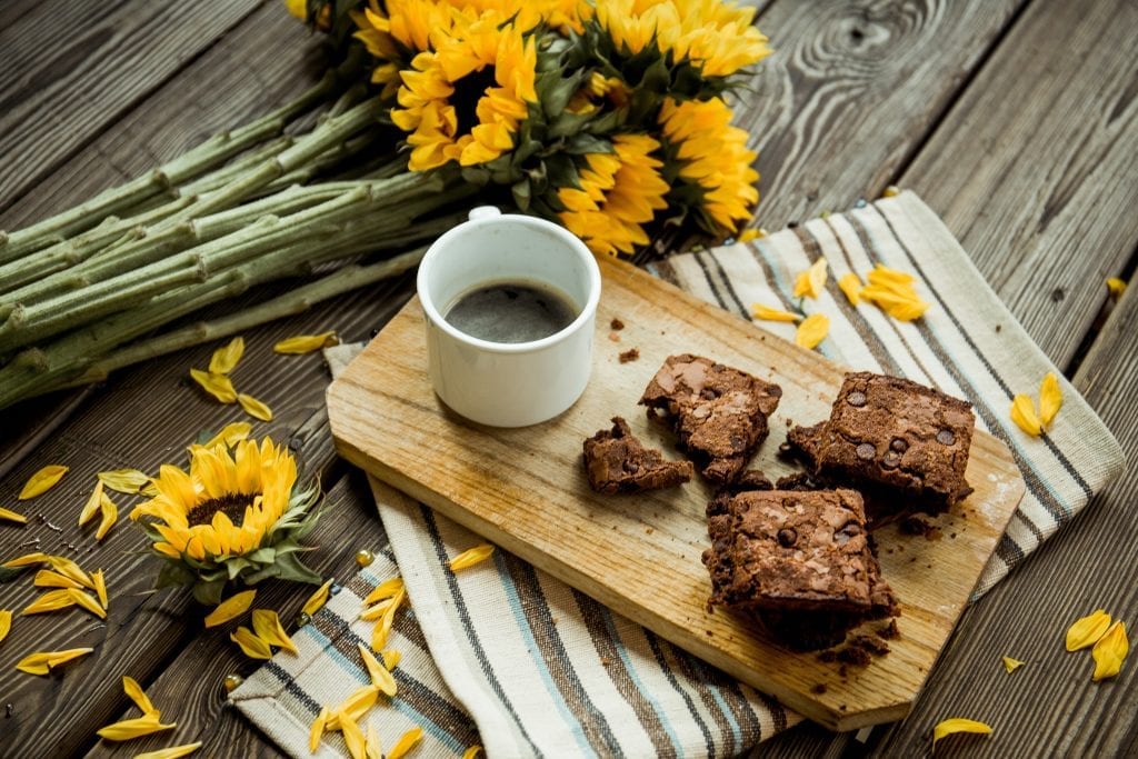 cannabis brownies and cup of coffee on a cutting board. The cutting board is on a table with sunflowers