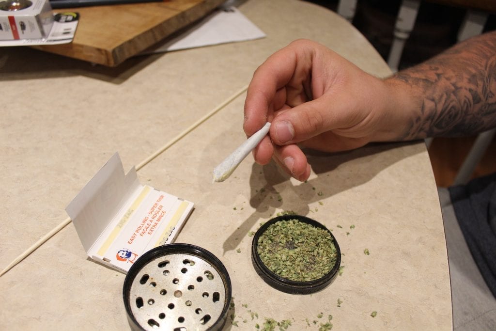 Perfectly rolled joint with cannabis in a grinder below it. Pack of rolling papers on the table beside the grinder.