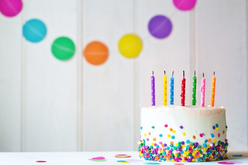 Cannabis birthday cake with sprinkles on the side of it with blown out candles in front of a wall with birthday decorations on it.
