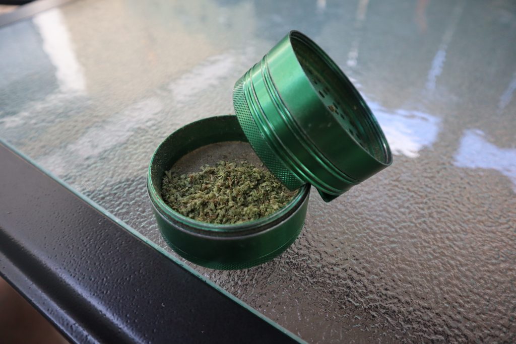 A green grinder that is opened is filled with ground up cannabis bud. The grinder sits on a glass table. 