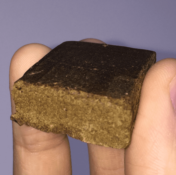 a man holds a brown square chunk of pressed hash in between his fingers. 